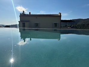 A clear swimming pool reflecting a building with a tiled roof and shutters, surrounded by greenery and hills.