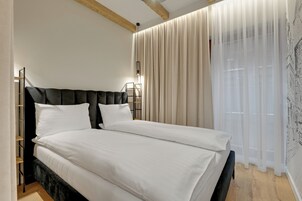 Egy franciaágyas ágy fehér ágyneművel, sötét színű ágyvégfallal és éjjeli lámpákkal.
