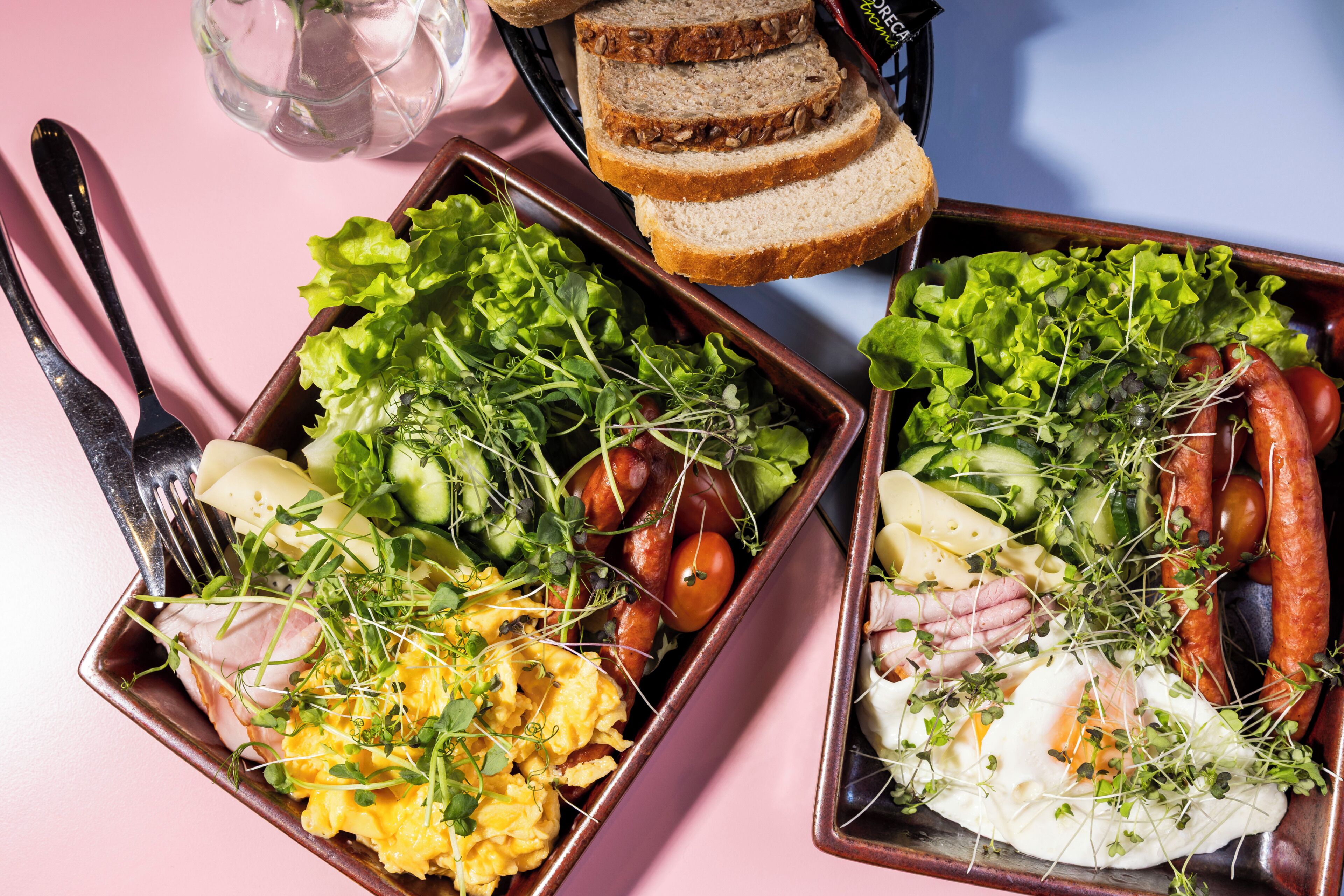 Two rectangular dishes with various food items, including vegetables, eggs, and sausages, on a pink surface.
