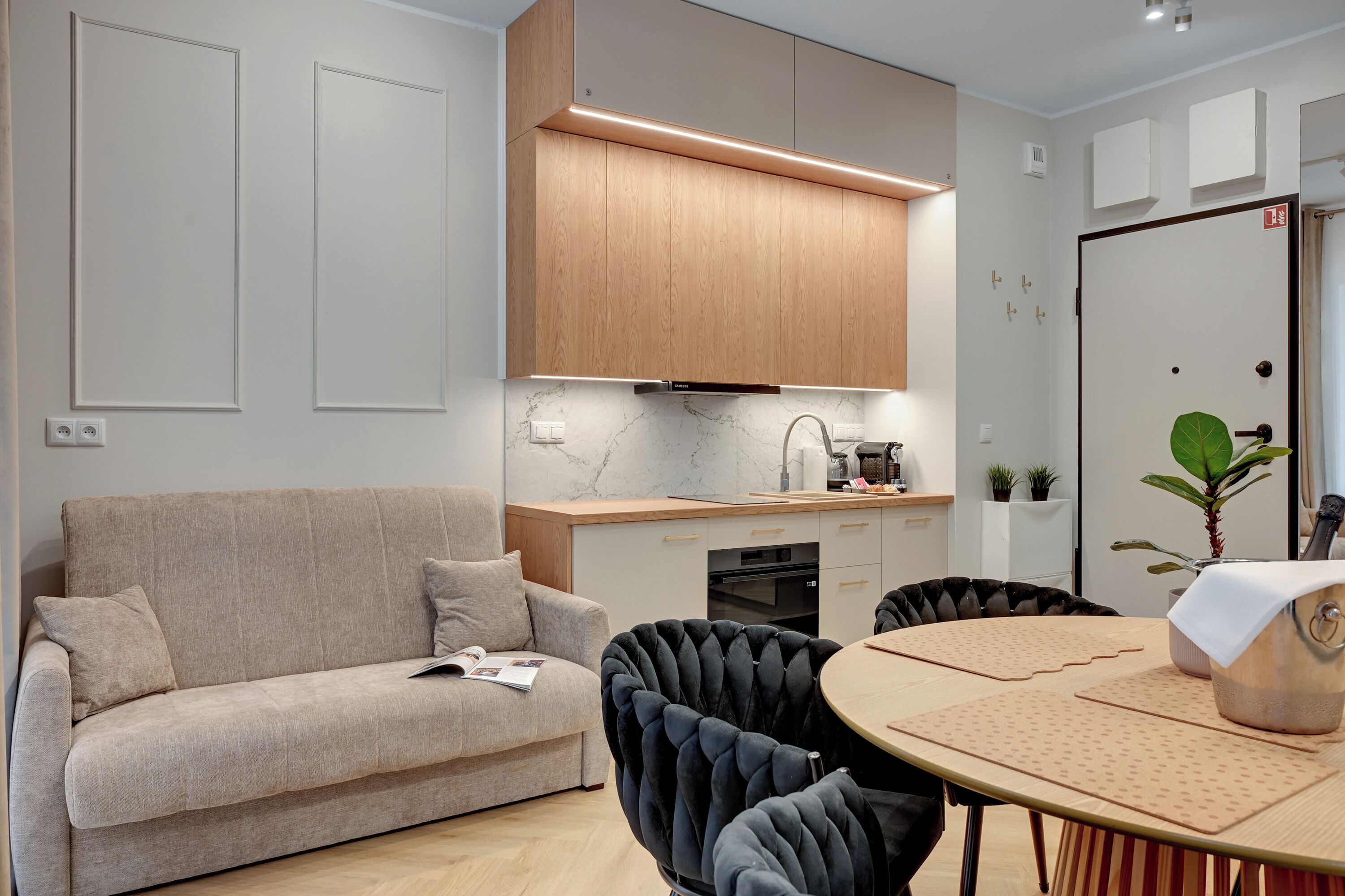 A modern living room with a sofa, a dining table with chairs, a kitchen area with wooden cabinets and a countertop, and a door with a plant.