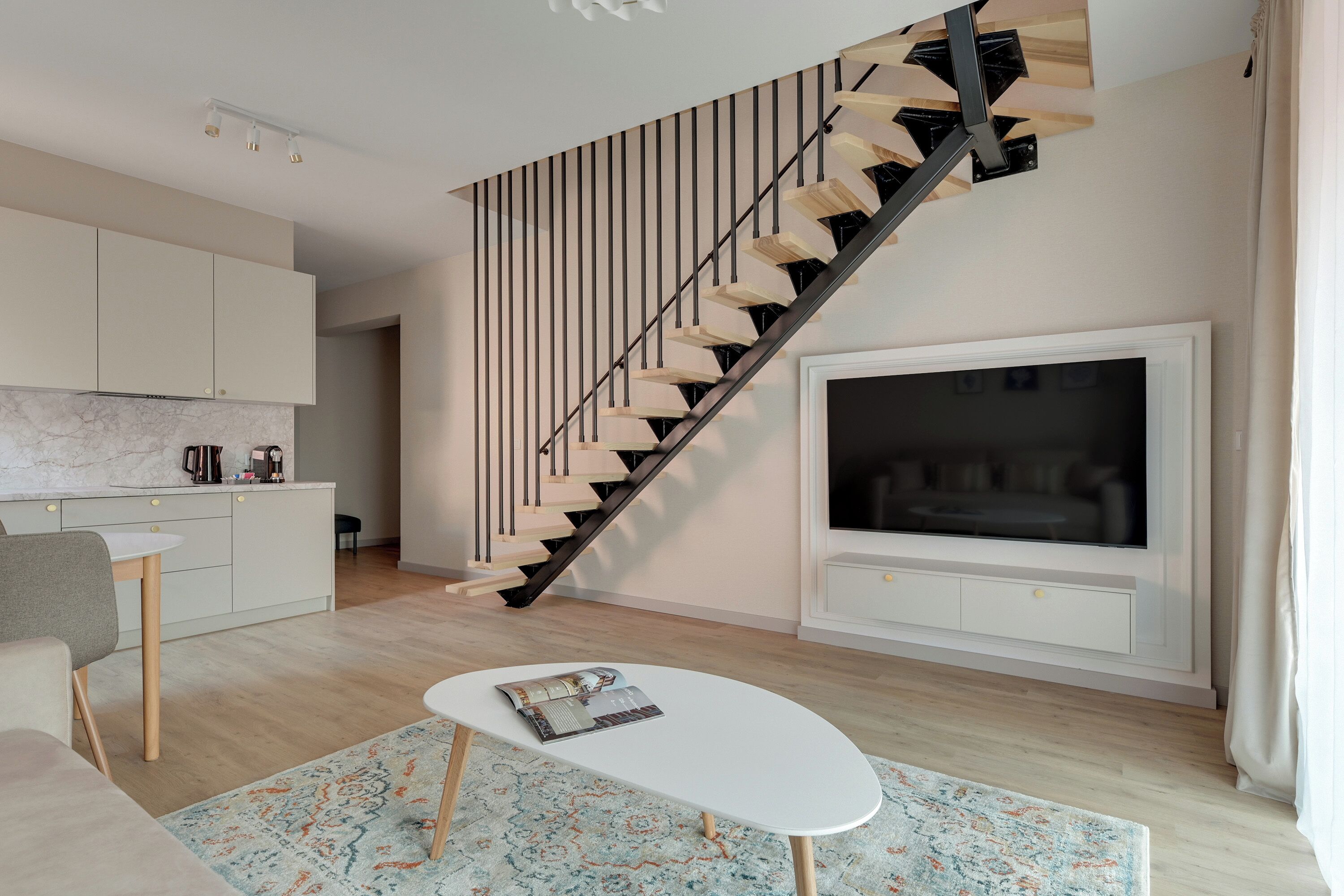 A modern living room with a staircase, a flat-screen TV, a white coffee table, and a kitchen area.