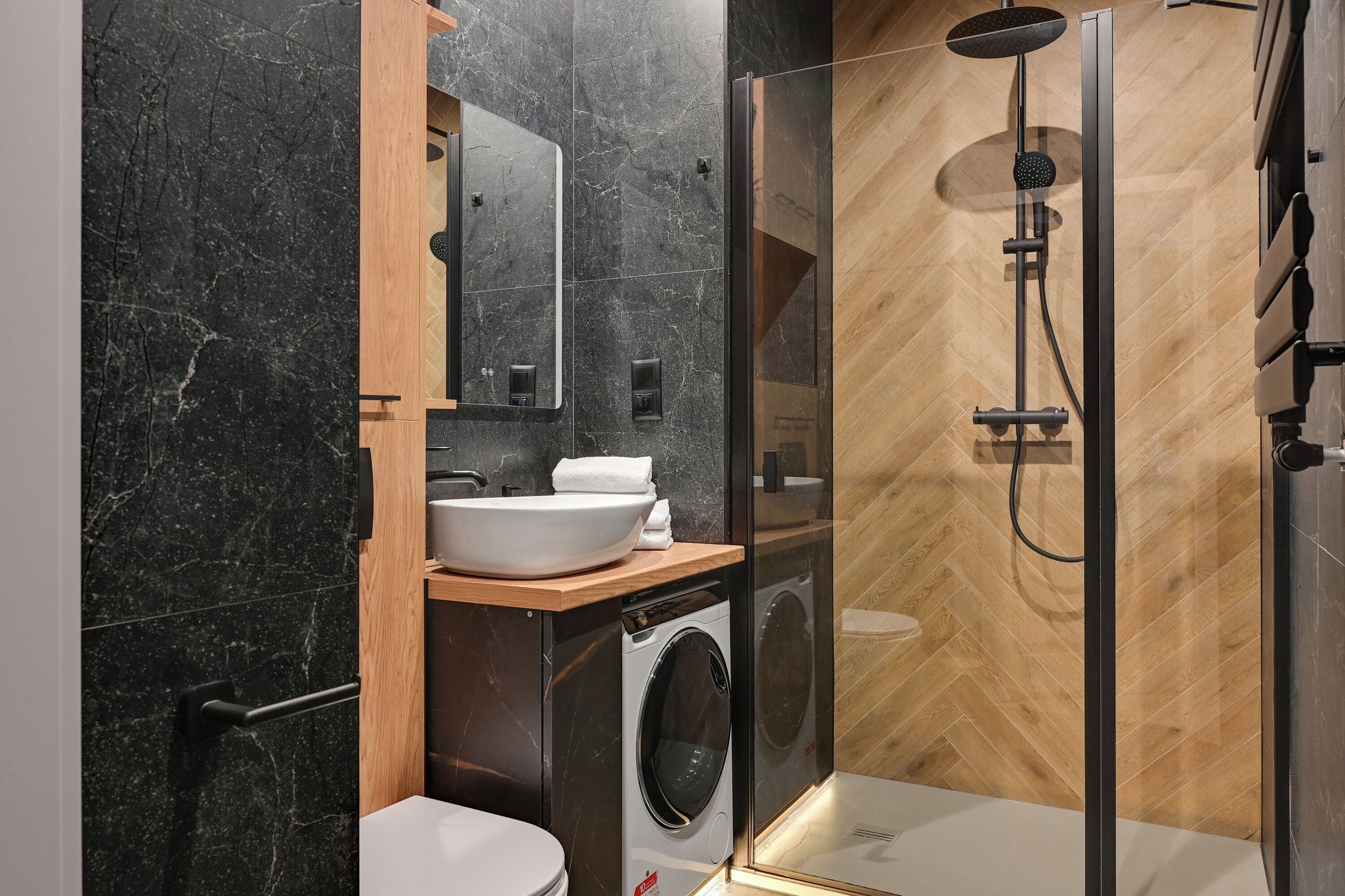A modern bathroom with a marble sink, a mirror, a wooden vanity, a washing machine, and a glass-enclosed shower area.
