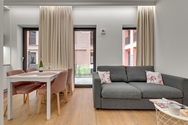 Una sala de estar moderna con un sofá gris, una mesa de comedor con sillas rosa y vistas a un edificio a través de grandes ventanales.