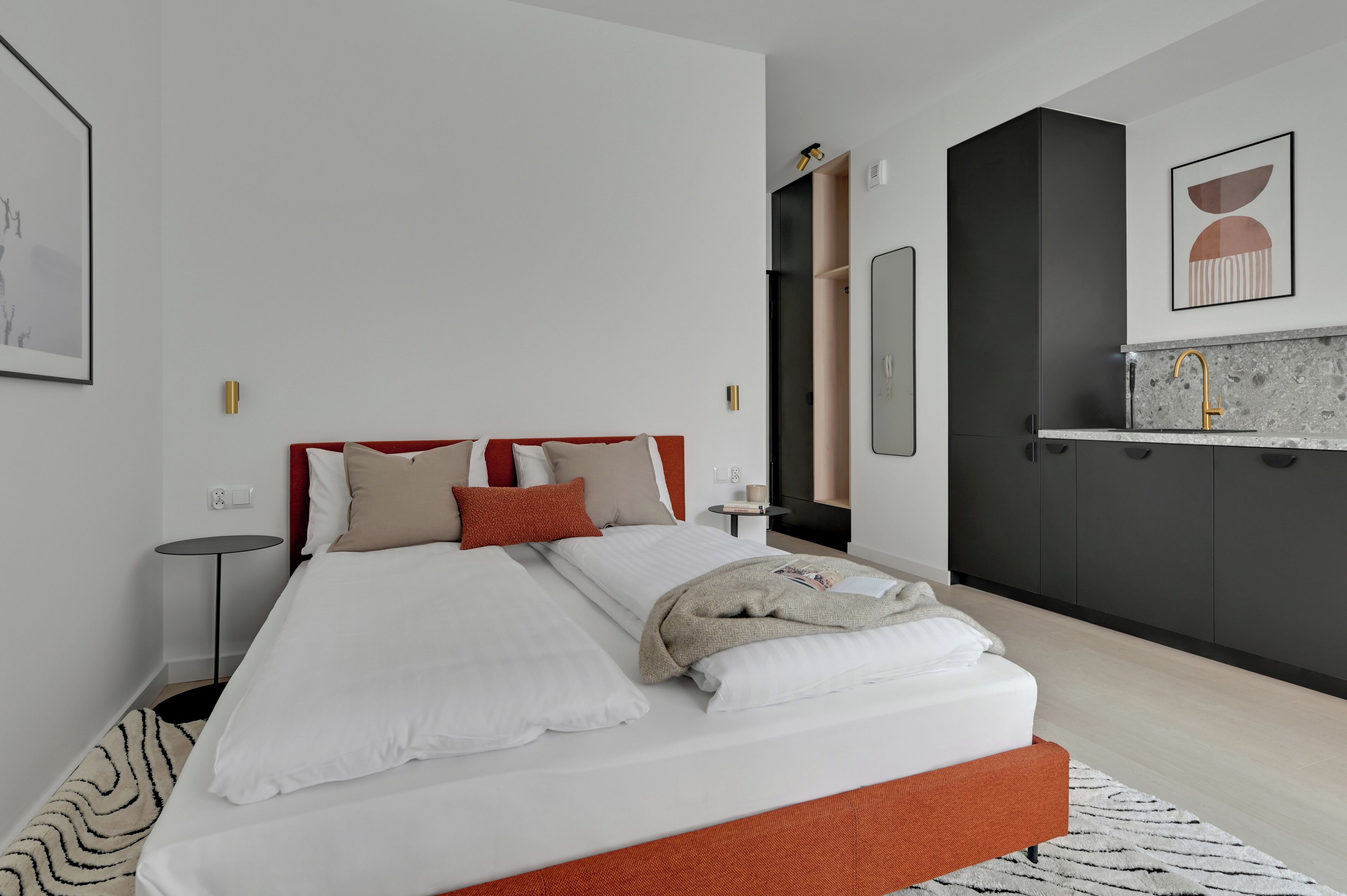 A modern hotel room with a large bed, a small bedside table, a built-in wardrobe, and a kitchenette area.