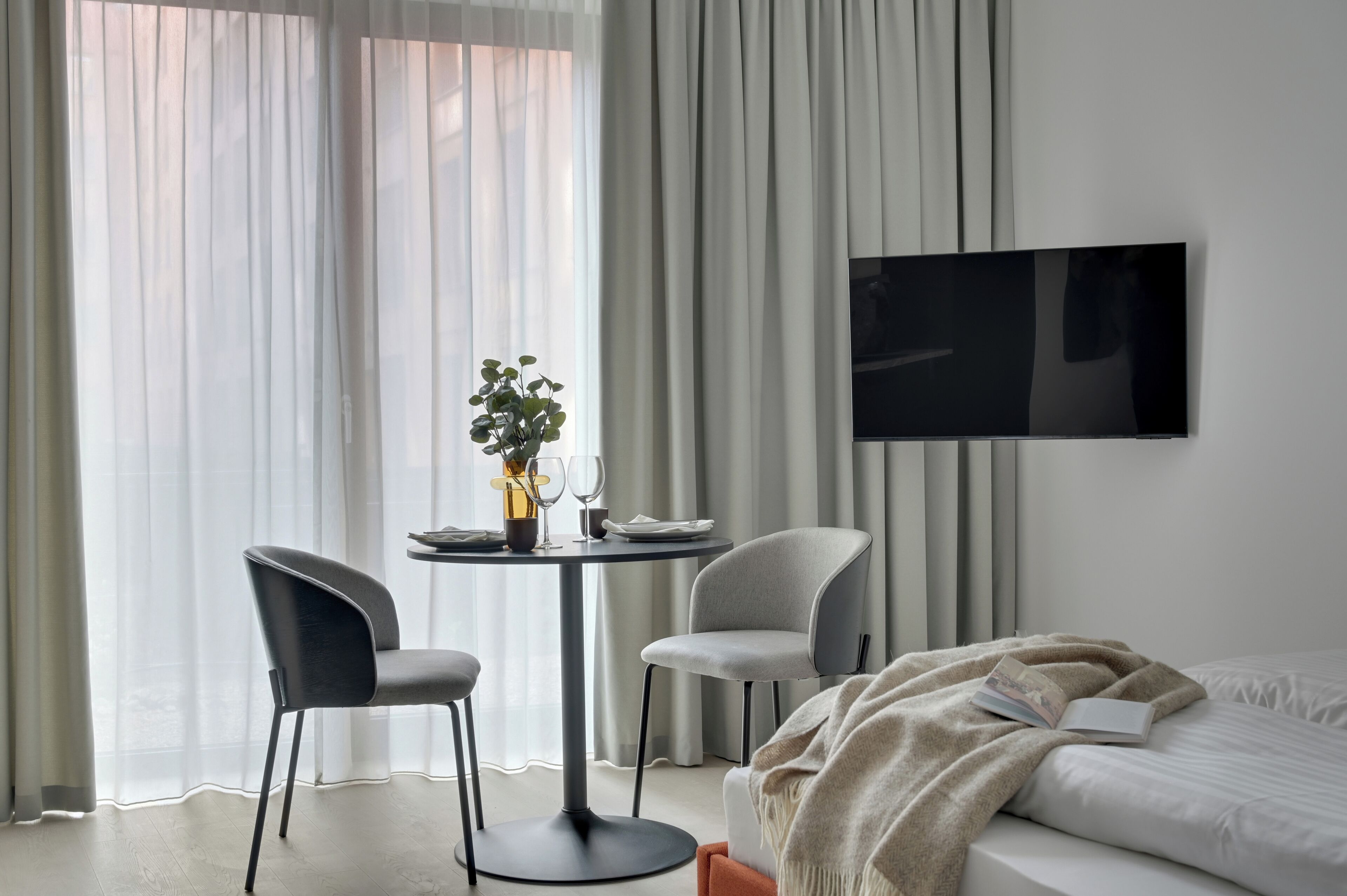 Ein modernes Hotelzimmer mit einem Bett, einem an der Wand befestigten Fernseher, einem Esstisch mit zwei Stühlen und einem großen Fenster mit durchsichtigen Vorhängen.
