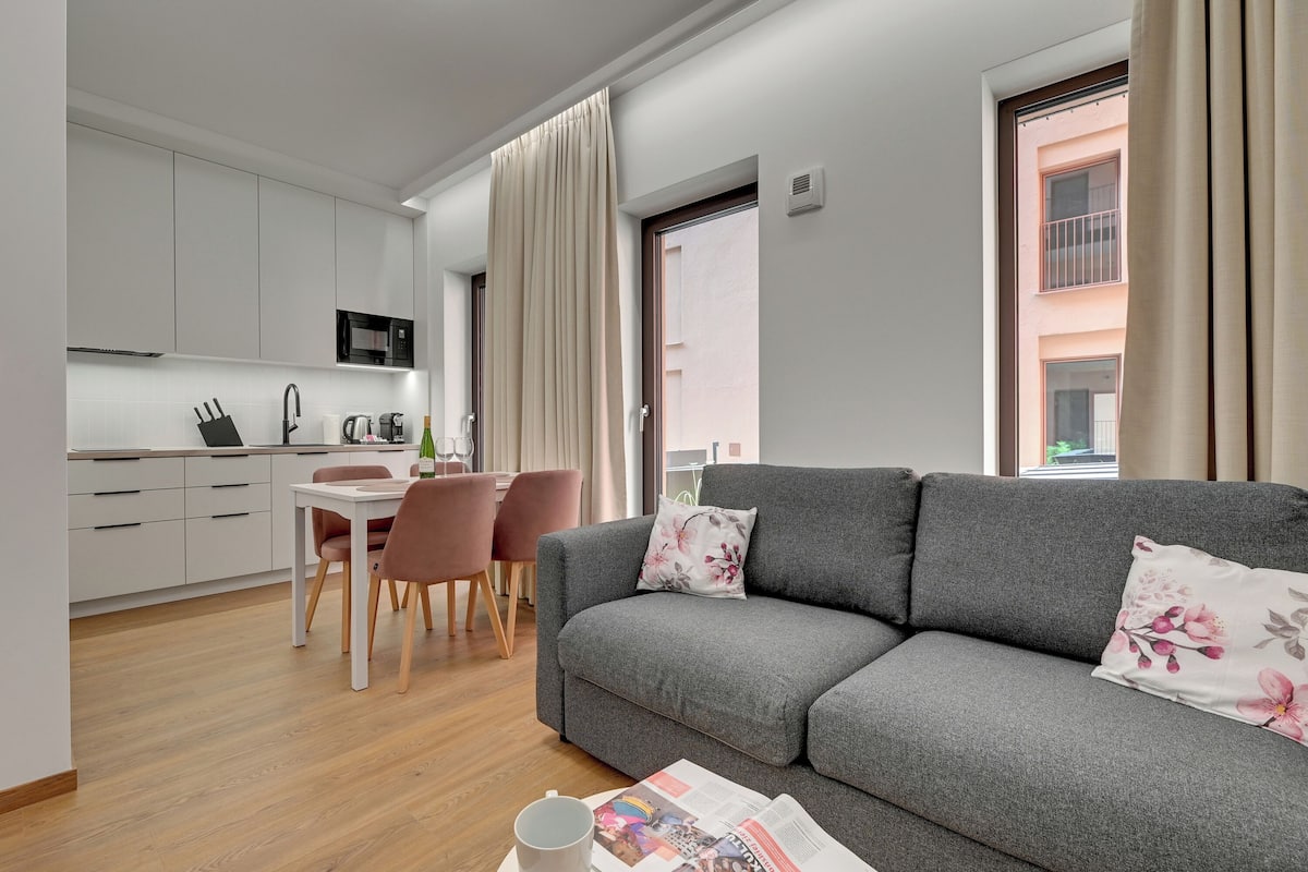Un soggiorno moderno con un divano grigio, un tavolo da pranzo con sedie, una zona cucina con mobili bianchi e una finestra con tende.
