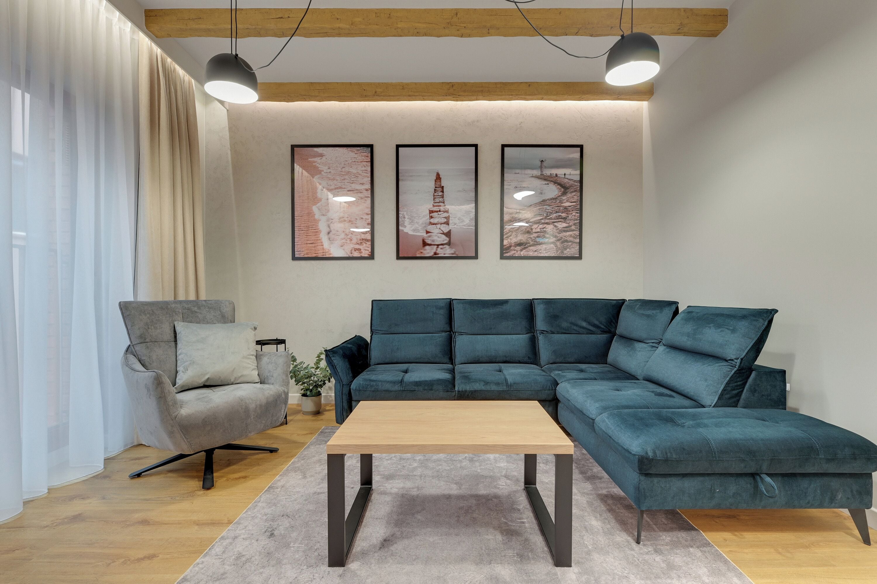 Ein modernes Wohnzimmer mit einem Ecksofa, einem Holztisch und einem grauen Sessel. An der Wand hängen drei gerahmte Bilder und auf dem Sofa steht ein Blumentopf.