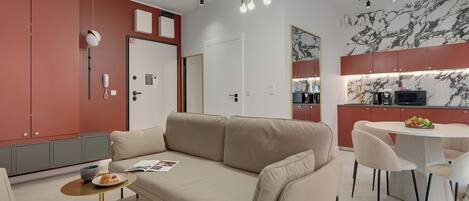 A modern living room with a beige sofa, a small round table with a plate of food, and a kitchen area with a marble backsplash and red cabinets.