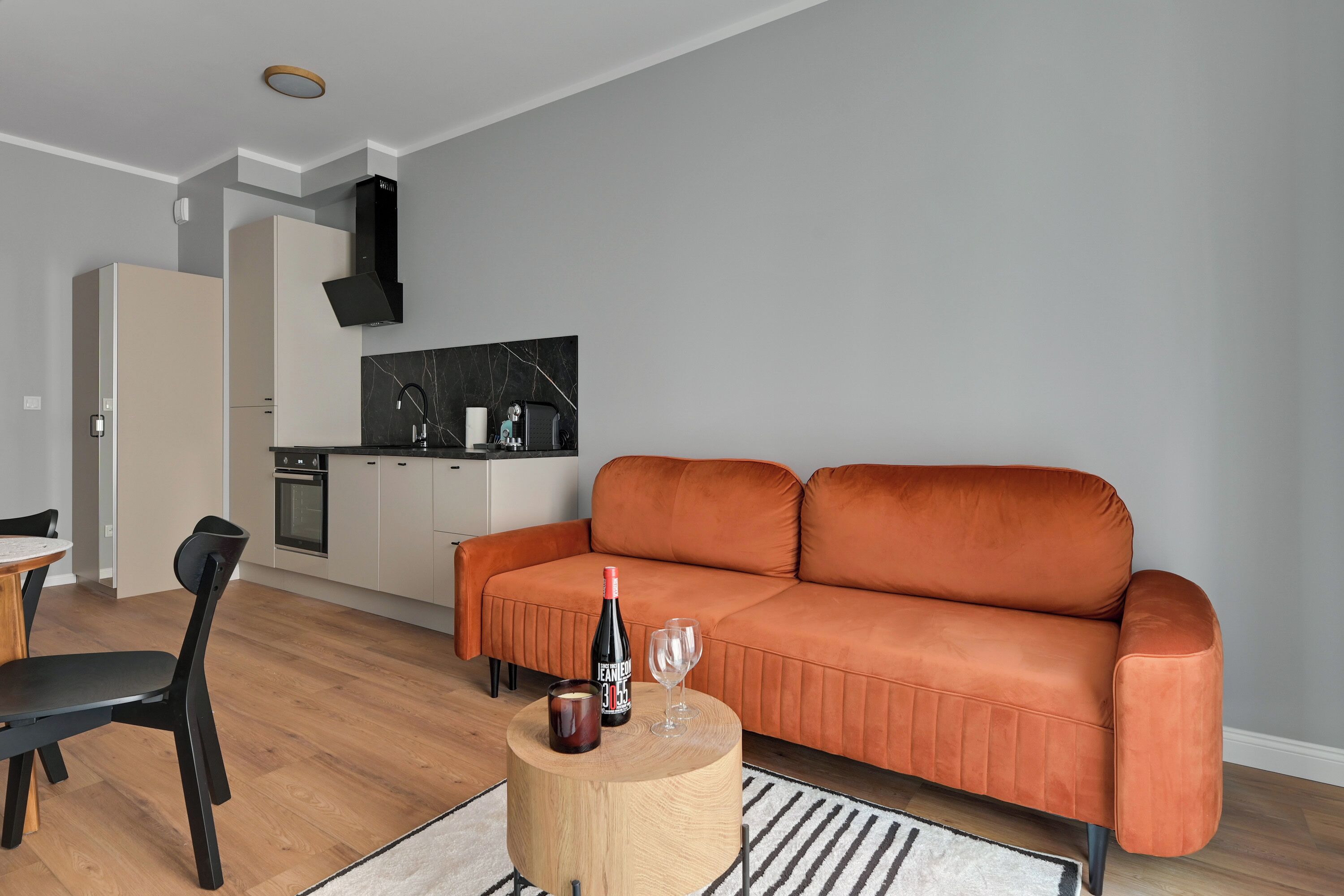 Una sala de estar moderna con un sofá, una mesa redonda de madera, una zona de comedor con una silla y una cocina al fondo.