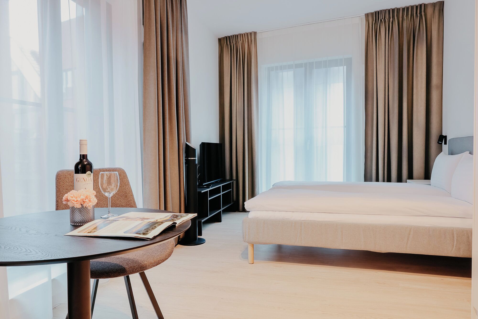 Hotellihuone, jossa on sänky, televisio, pöytä, jonka päällä on viinipullo ja lasit, sekä verhot.
