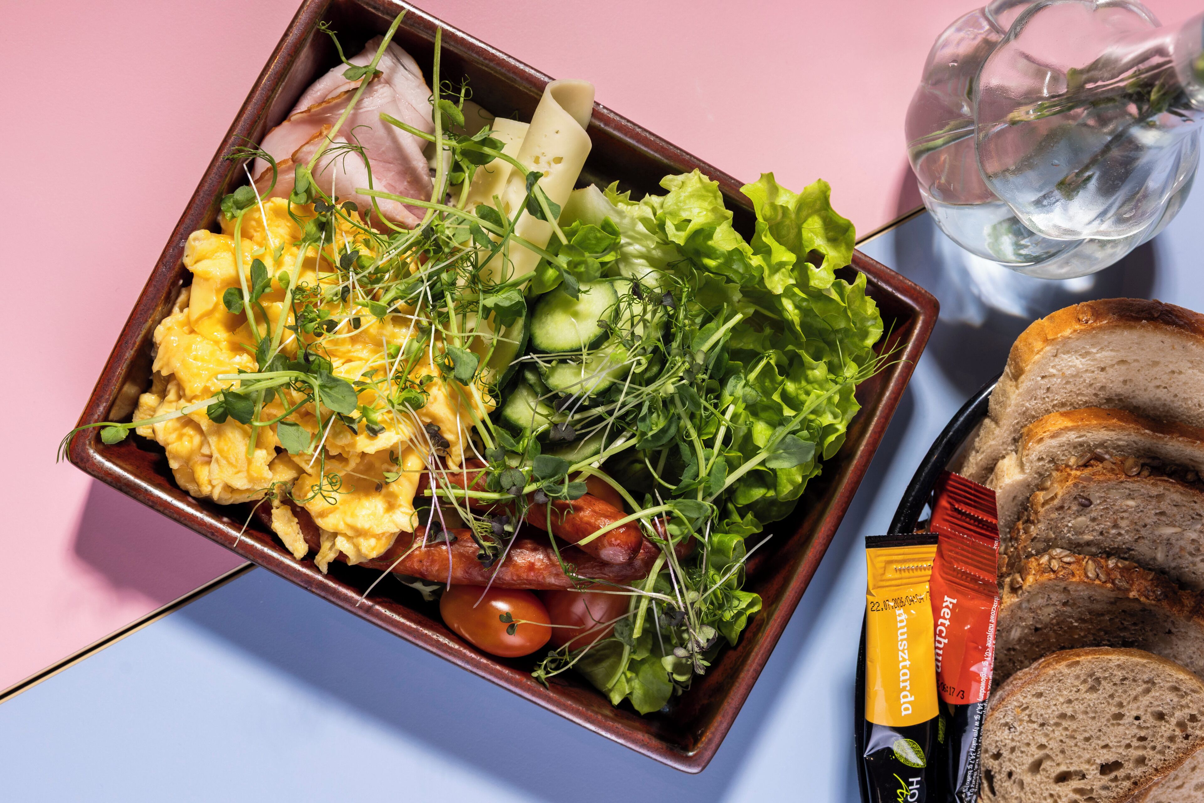 A meal with scrambled eggs, sausages, and vegetables in a ceramic dish, accompanied by sliced bread and a glass of water on a table.