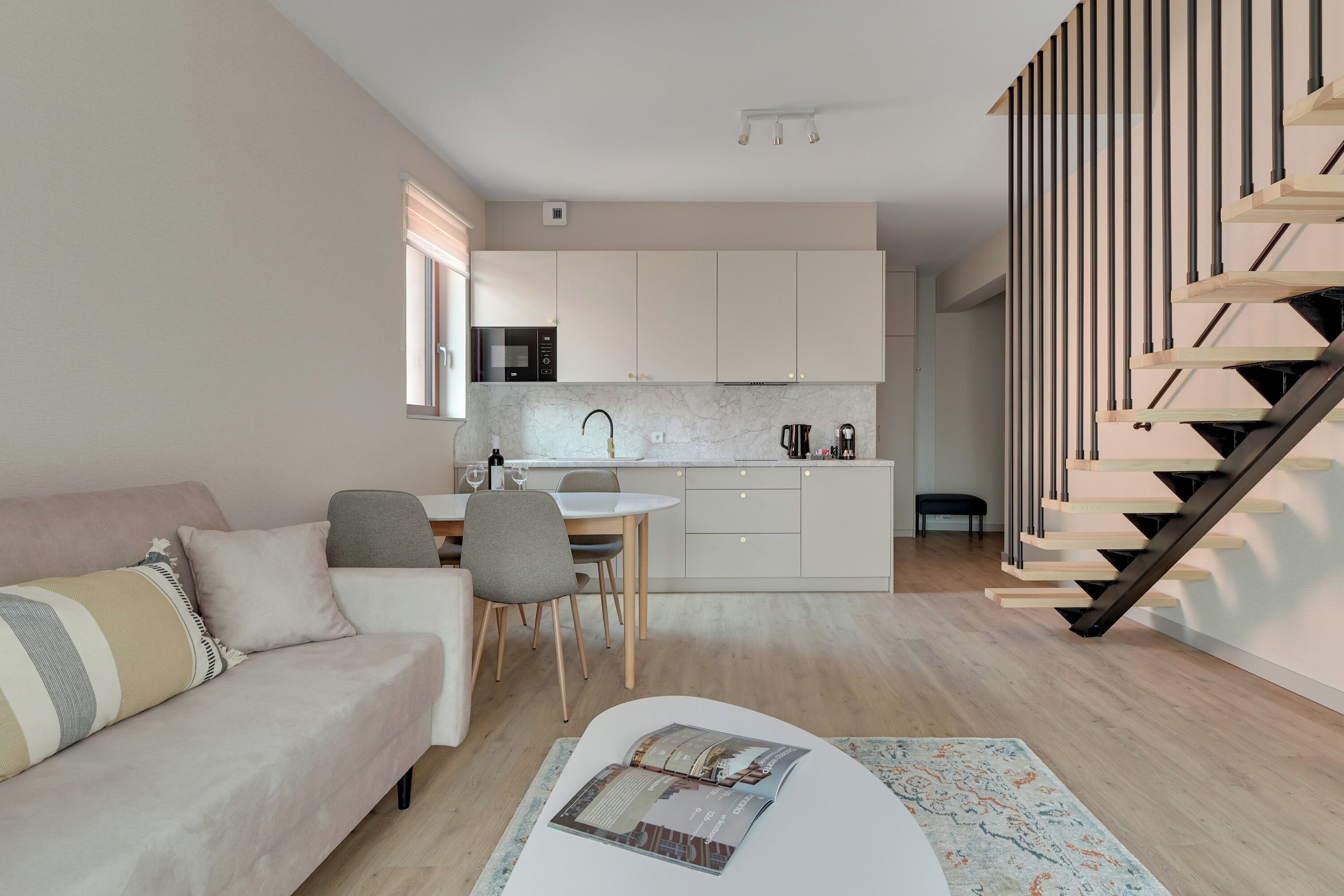 Une cuisine moderne avec des meubles blancs, un coin repas comprenant une table et des chaises, un canapé et un escalier avec des marches en bois.
