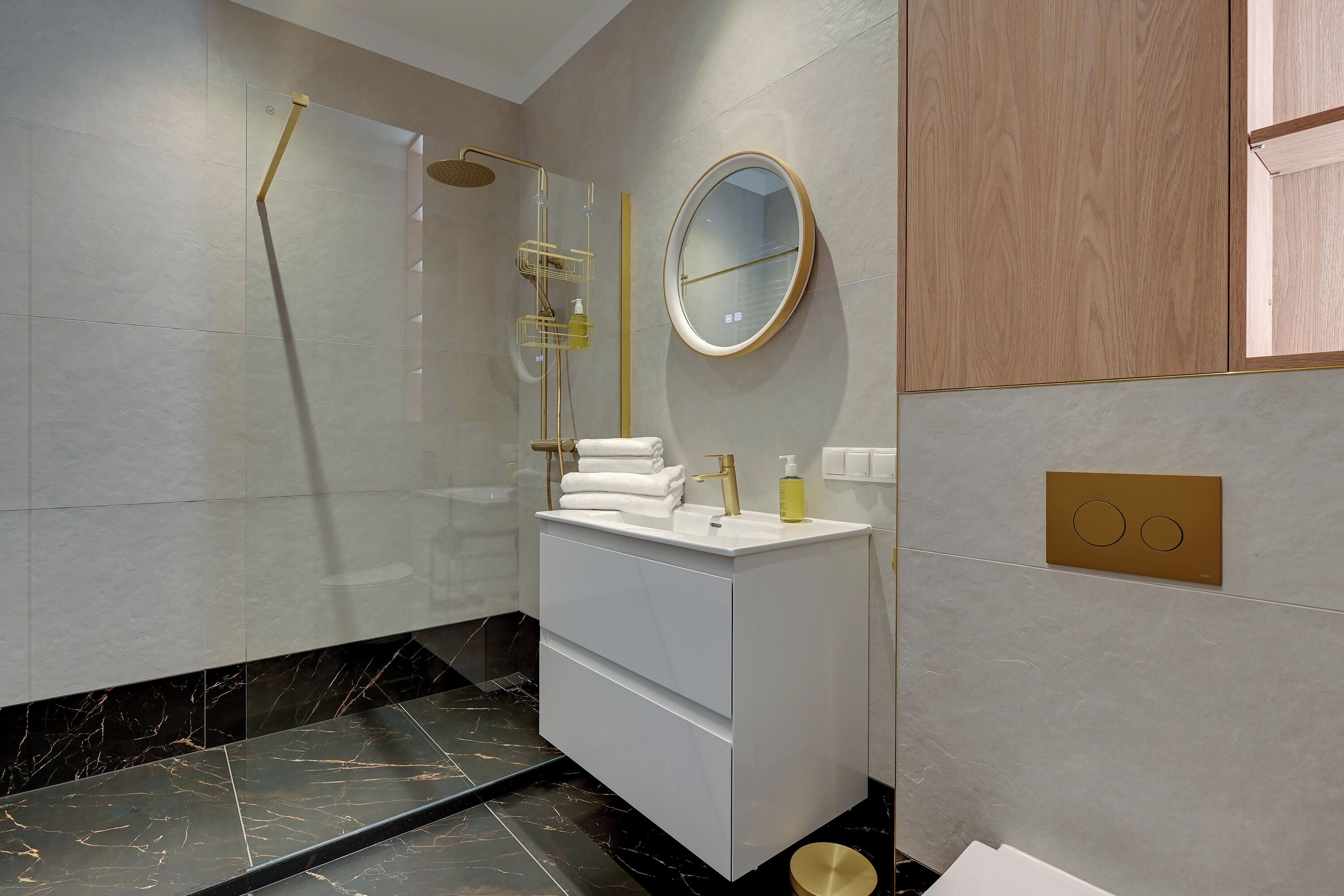 Un baño moderno con ducha de vidrio, tocador blanco con grifo dorado y espejo redondo en la pared.