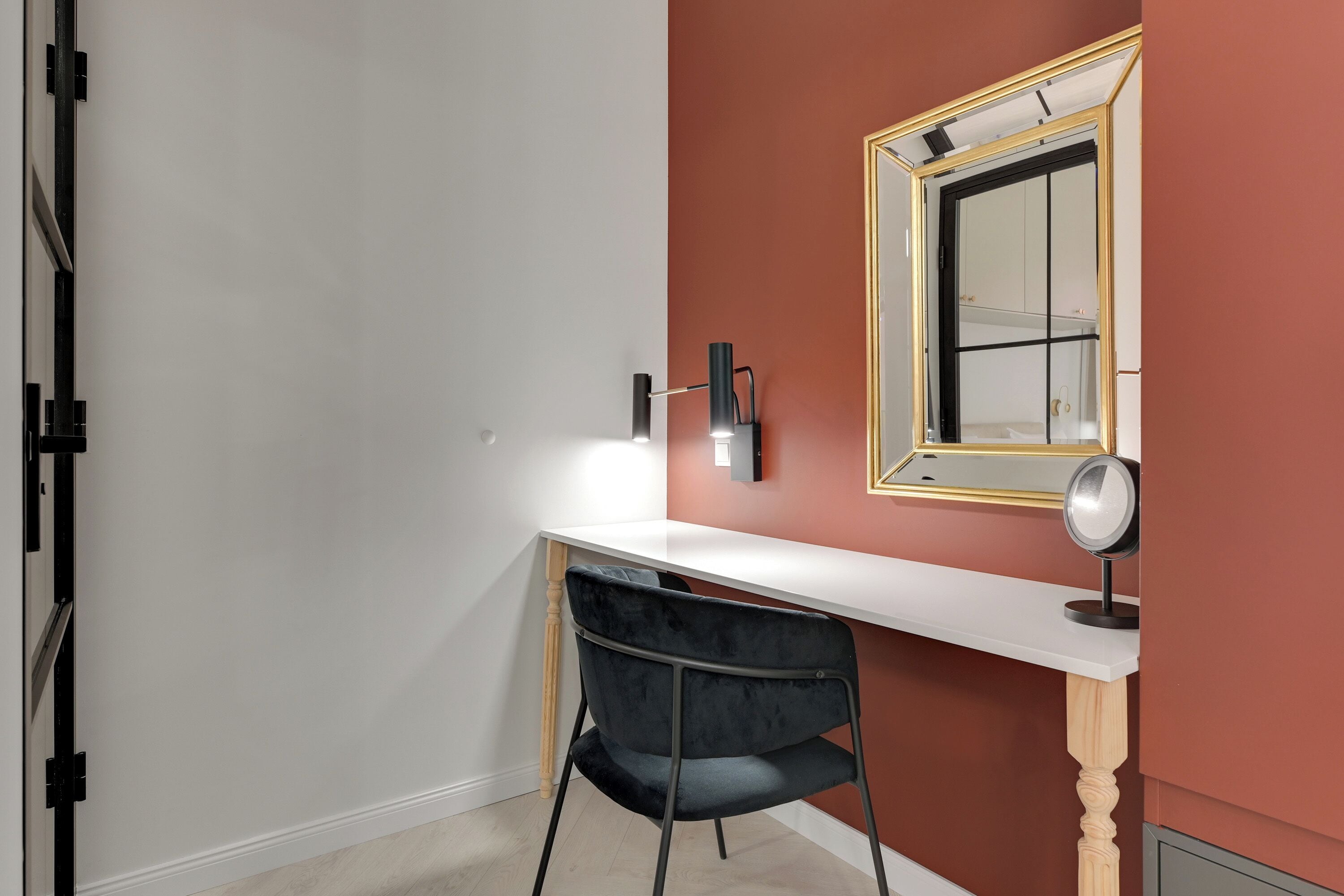 A room with a white desk, a black chair, a mirror with a gold frame, and a wall-mounted lamp.