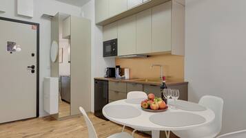 A modern kitchen with white cabinets, a microwave, and a sink. There is a dining area with a white table and chairs, and a bowl of fruit.