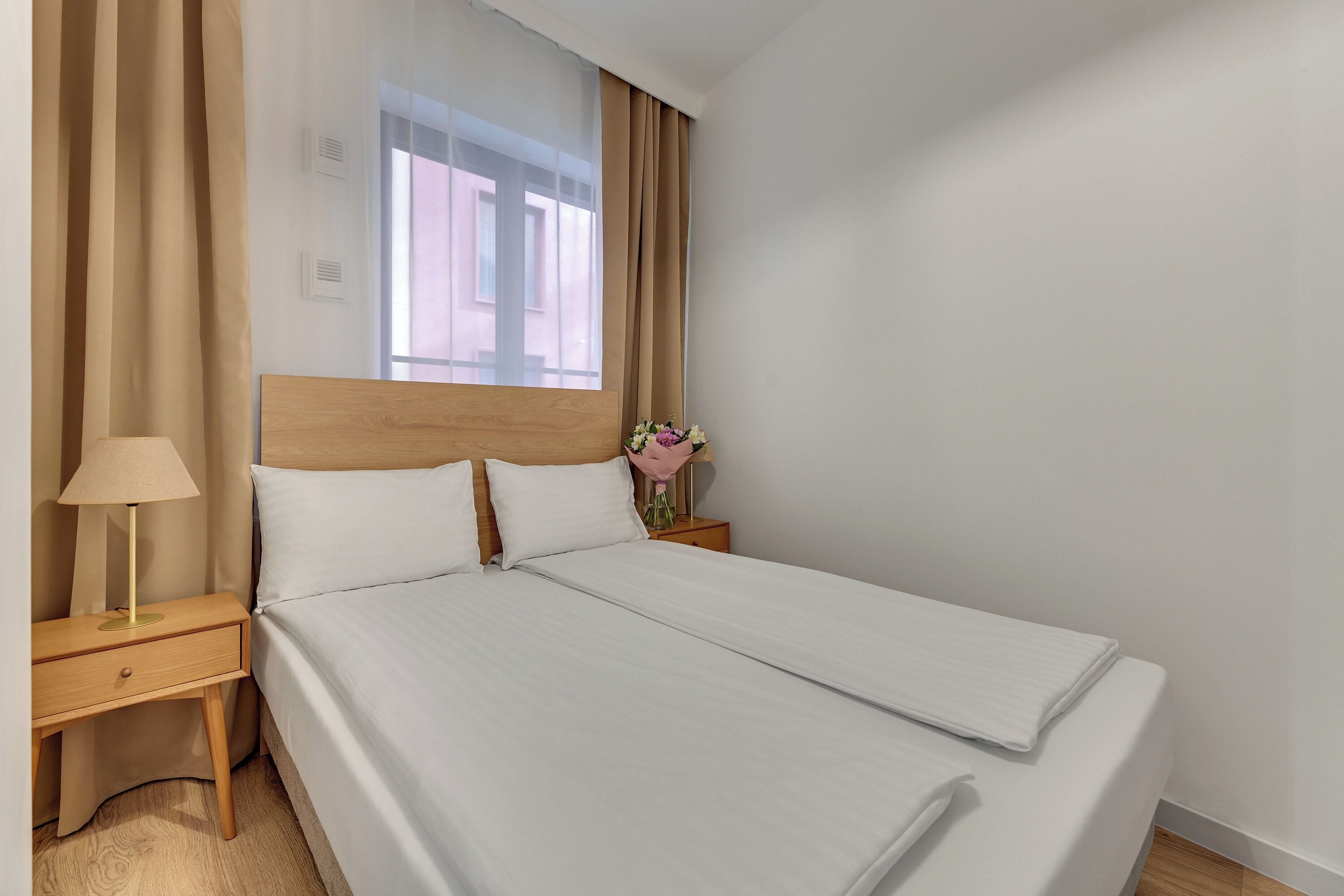 Una cama doble con ropa blanca, cabecera de madera, mesita de noche con lámpara y ventana con cortinas beige.