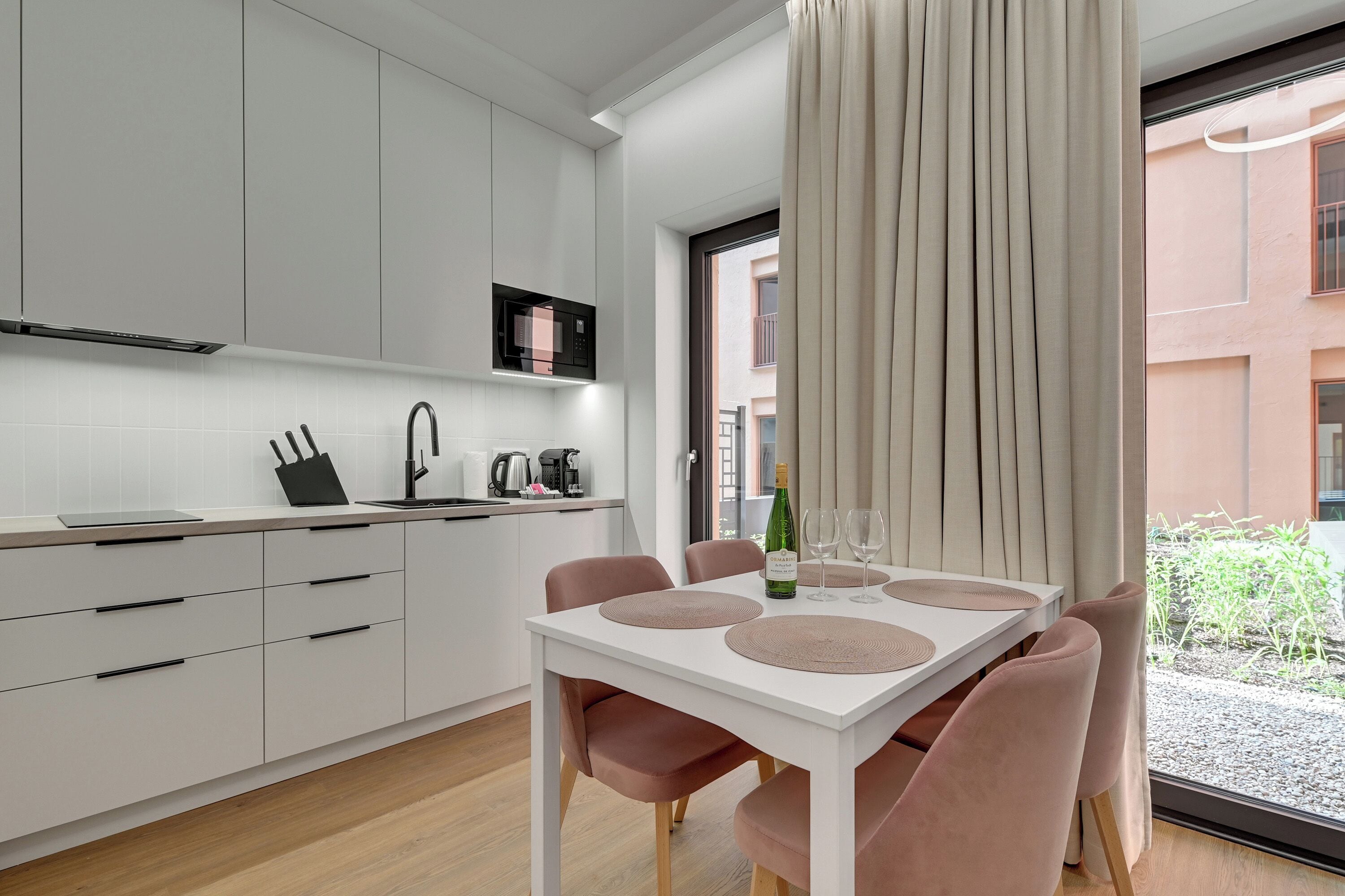A modern kitchen with white cabinets, a white dining table with pink chairs, and a view of a building through a window.