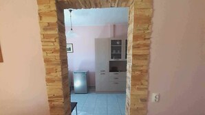 A room with a stone archway leading to a kitchen area with cabinets and a refrigerator. - The Cottage in Paralia (Paralia)