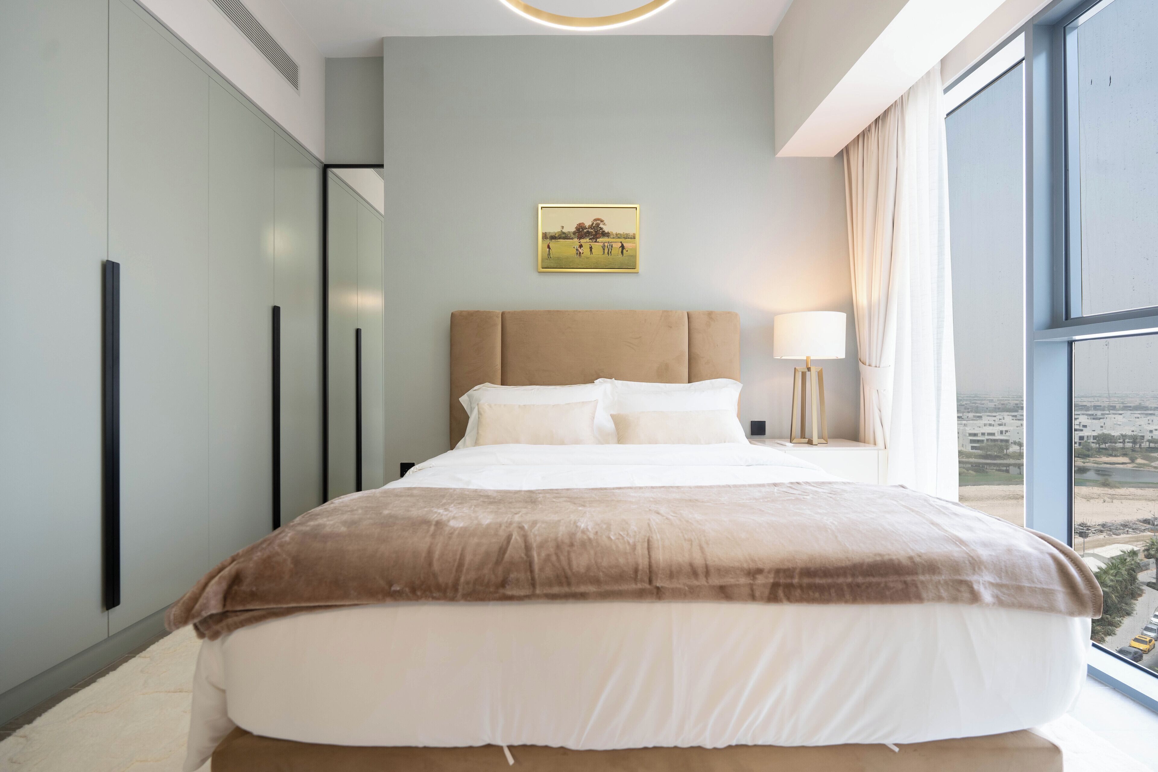 A modern bedroom with a large bed, a bedside lamp, and a view of a cityscape through a large window.