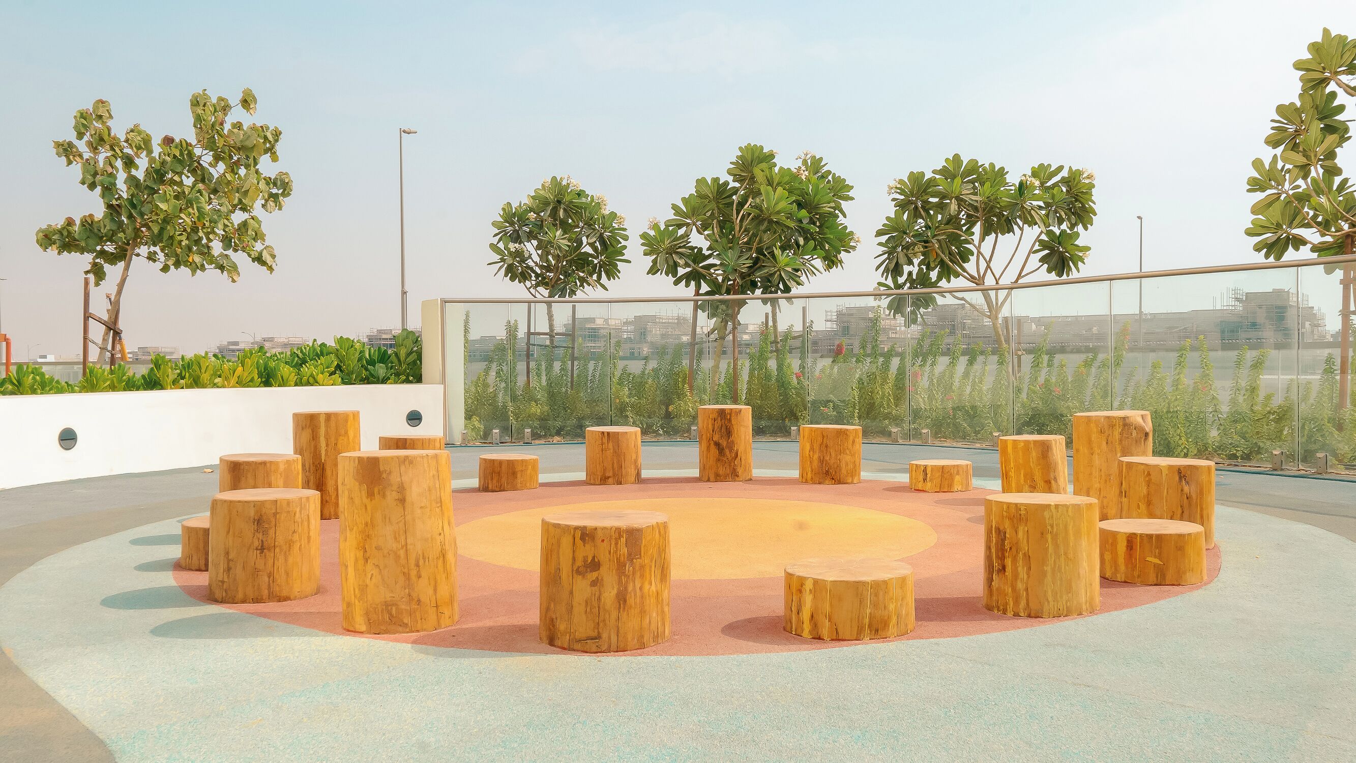 A circular seating area with wooden stools arranged around a central space, surrounded by greenery and a glass barrier.