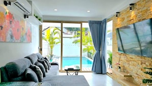 A modern living room with a grey sofa, a flat-screen TV mounted on a stone wall, a pool visible through the glass doors, and a colorful abstract painting on the wall. - Lux Sila Pool Villa 3 Bedrooms 4 Beds (Koh Samui)