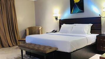 Habitación de hotel con una cama grande, una mesita de noche, una silla, un banco, un teléfono en la pared y un cuadro.