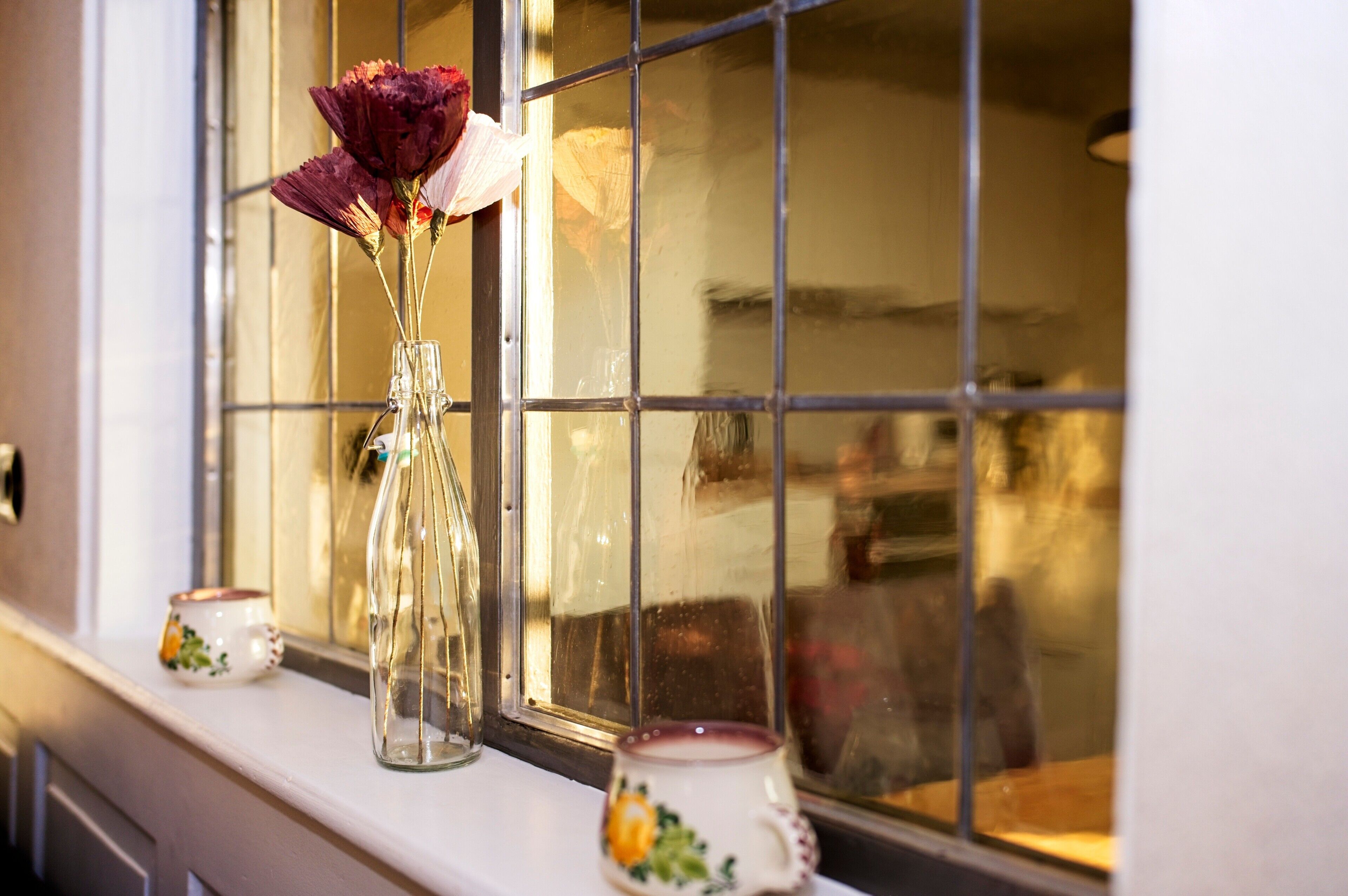 Ein Fenster mit freiem Blick in einen Raum, eine Vase mit Blumen auf einem Fensterbrett und eine Tasse mit floralem Dekor.