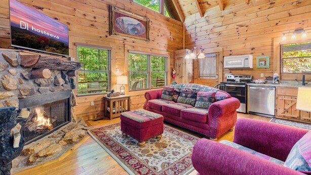 A cozy log cabin interior with a stone fireplace, a red sofa, and a kitchen area. - Mountain Oasis (Sevierville)