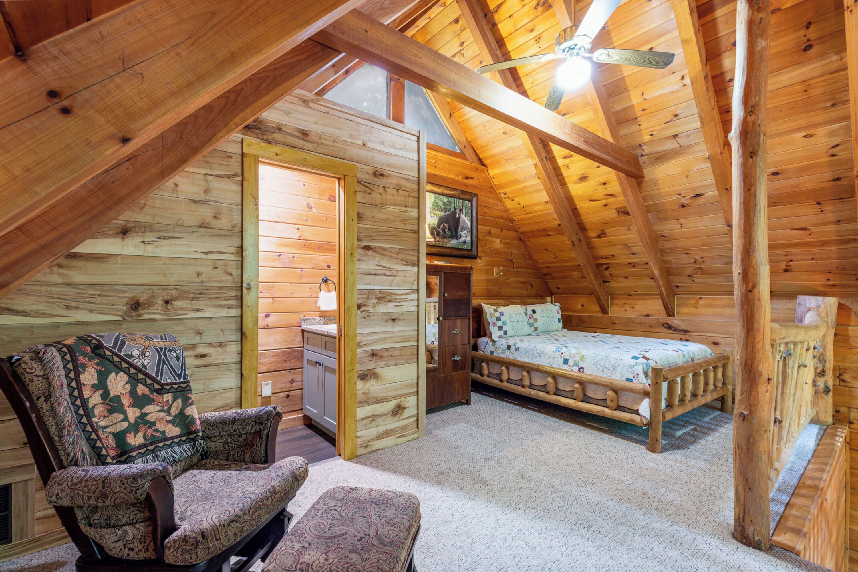 A cozy attic bedroom with a wooden ceiling, a bed, two chairs, and a small desk.