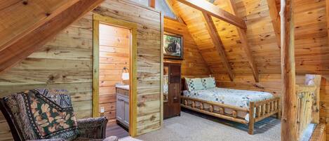 A cozy attic bedroom with a wooden ceiling, a bed, two chairs, and a small desk.