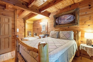 A wooden cabin bedroom with a bed, a nightstand, a lamp, a framed picture on the wall, and a bathroom visible through an open door.