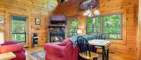 A cozy cabin interior with a fireplace, red sofa, dining table, and large windows offering a view of the outdoors.
