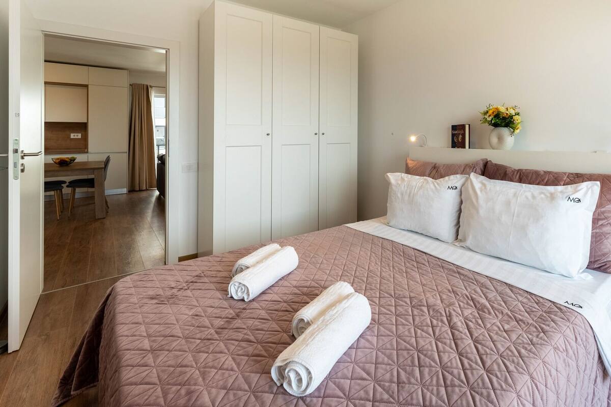 A bedroom with a large bed, two rolled towels, a headboard with multiple pillows, and a view into the adjacent dining area.