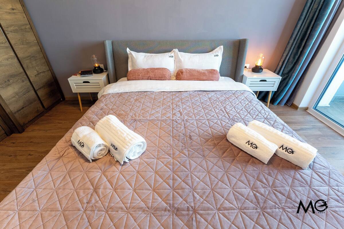 A hotel room with a large bed, two bedside tables, a headboard, and a window with curtains.