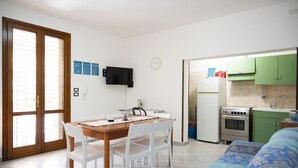 En kompakt opholdsstue med spisebord, stole, sofa, et tekøkken med komfur og køleskab samt et fjernsyn monteret på væggen.