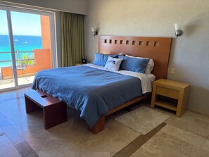 Una habitación de hotel moderna con una cama grande, una mesita de noche de madera, una pequeña mesa y vista al océano a través de una puerta corrediza.