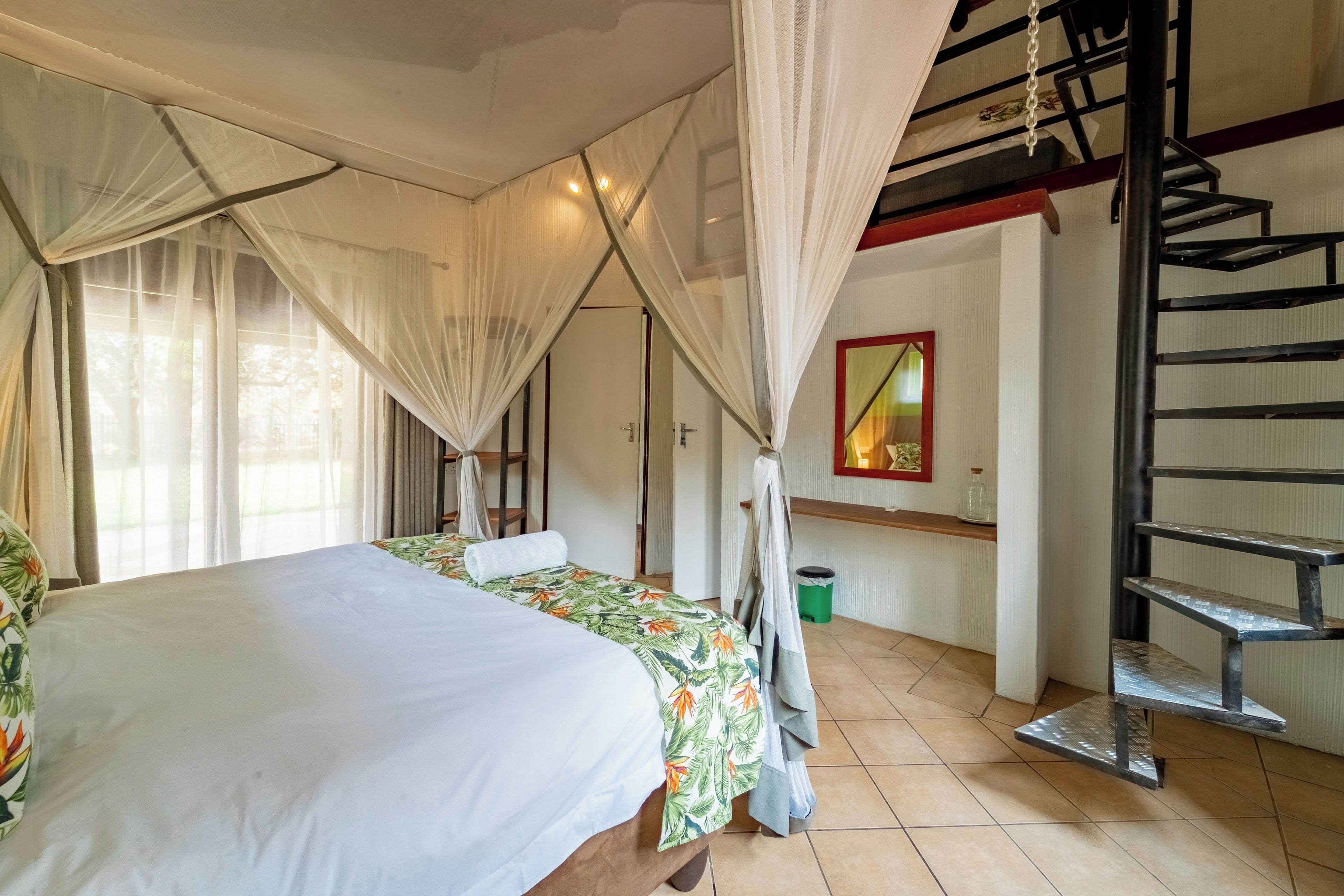 Ein Schlafzimmer mit einem Bett, einem Fenster mit durchscheinenden Vorhängen, einer Treppe und einem kleinen Regal.