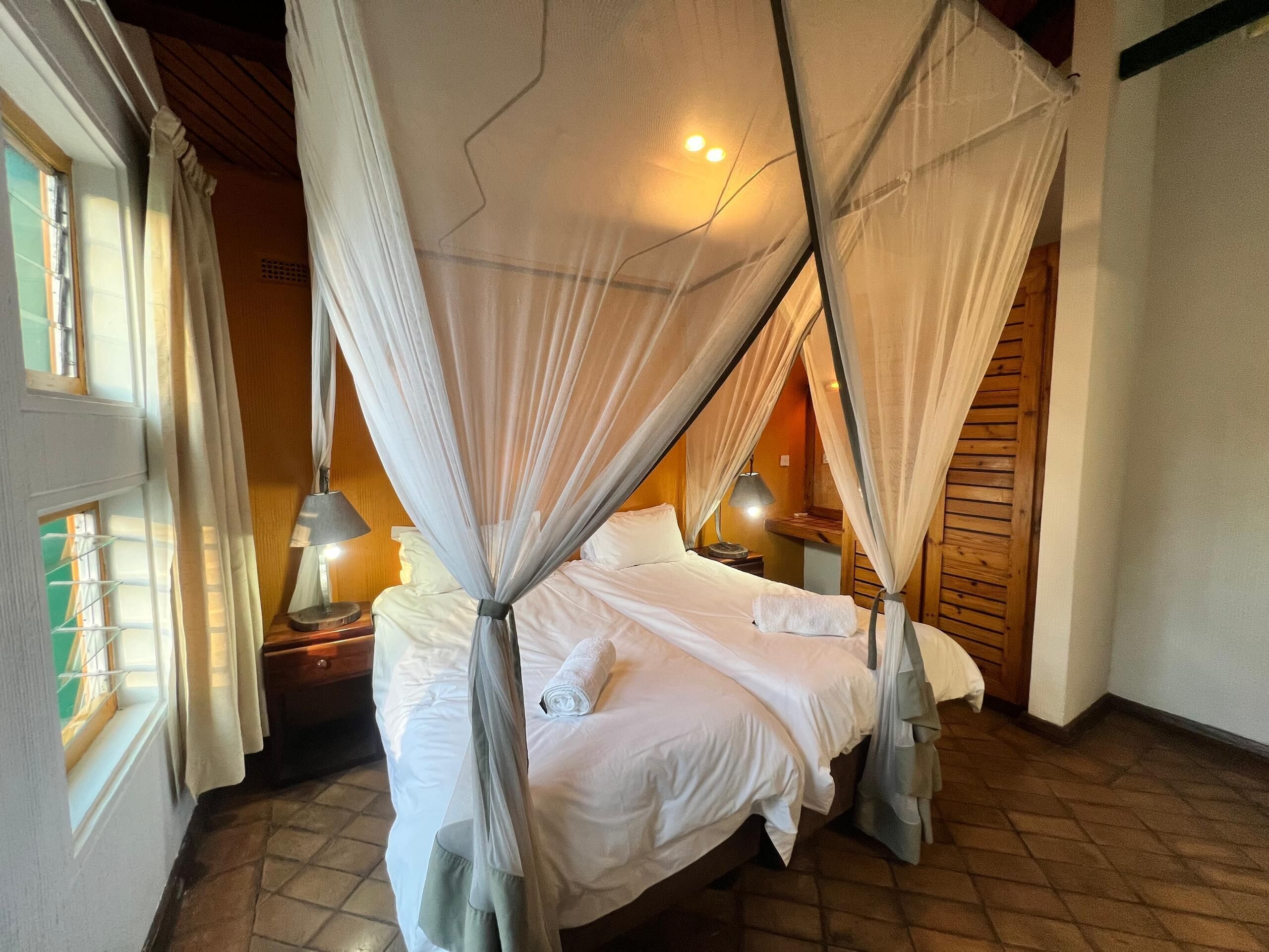 Una cama con dosel, sábanas blancas, una mesita de noche con lámpara y una ventana con cortinas.