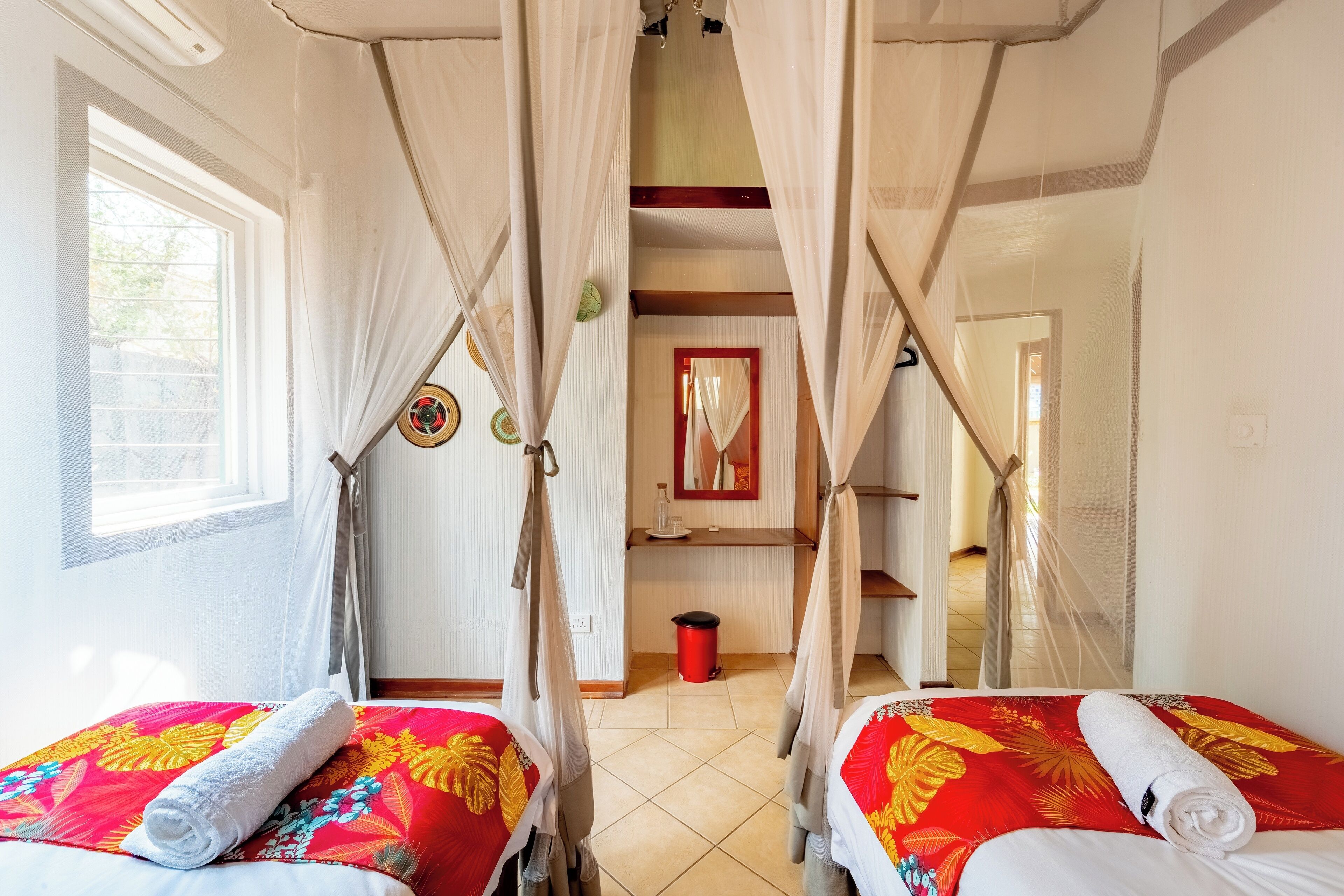 A hotel room with two beds, each with a red floral bedspread and white pillows. There is a window on the left side, a mirror on the wall, and a door leading to another room.