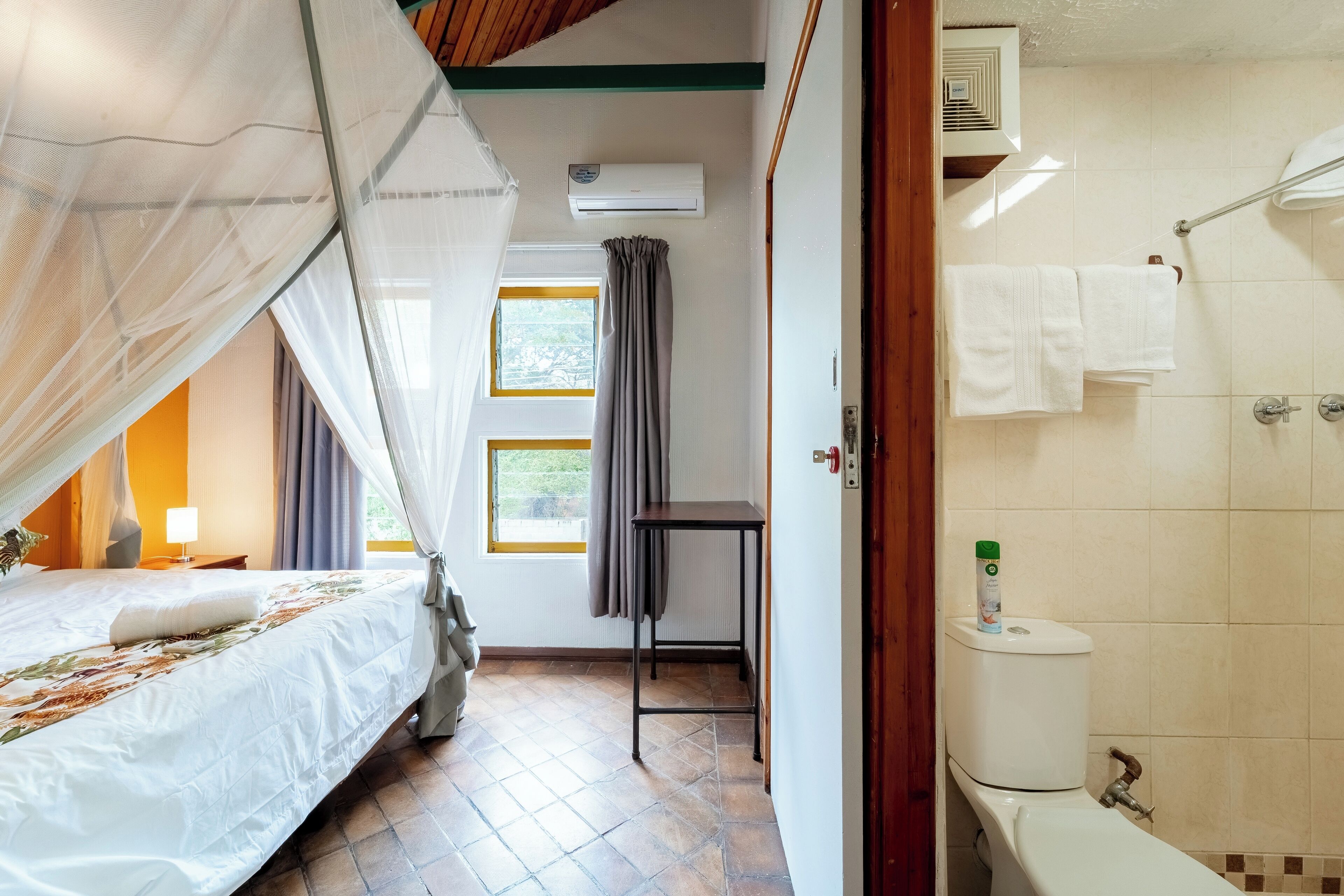 Ein Schlafzimmer mit Bett, Nachttisch, einem Fenster mit Vorhängen, einer Klimaanlage und Blick ins Freie.
