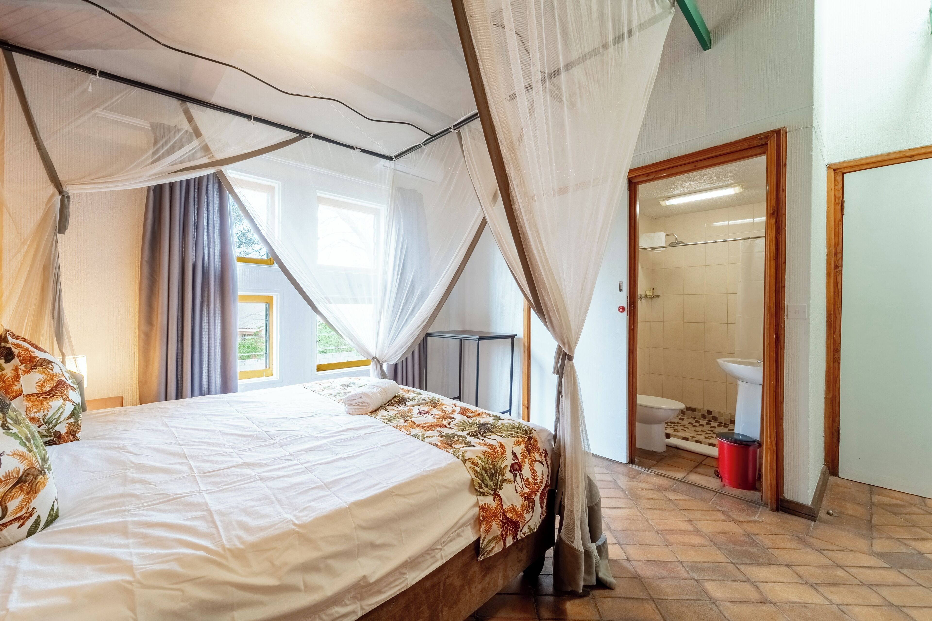 Ein Schlafzimmer mit Bett, einem Fenster mit Vorhängen, einem Badezimmer mit Toilette und Waschbecken sowie einer Duschkabine.