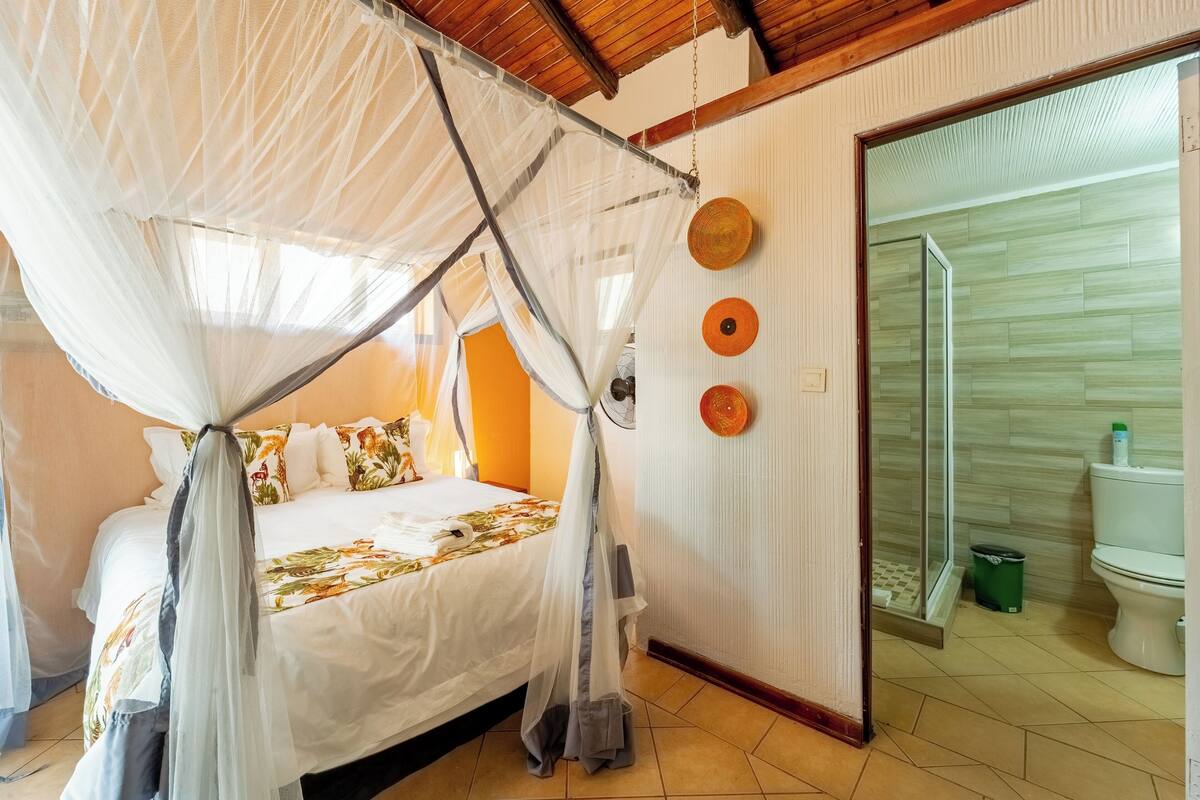 A bedroom with a bed under a mosquito net, a bathroom with a toilet and shower, and a door leading to another room.