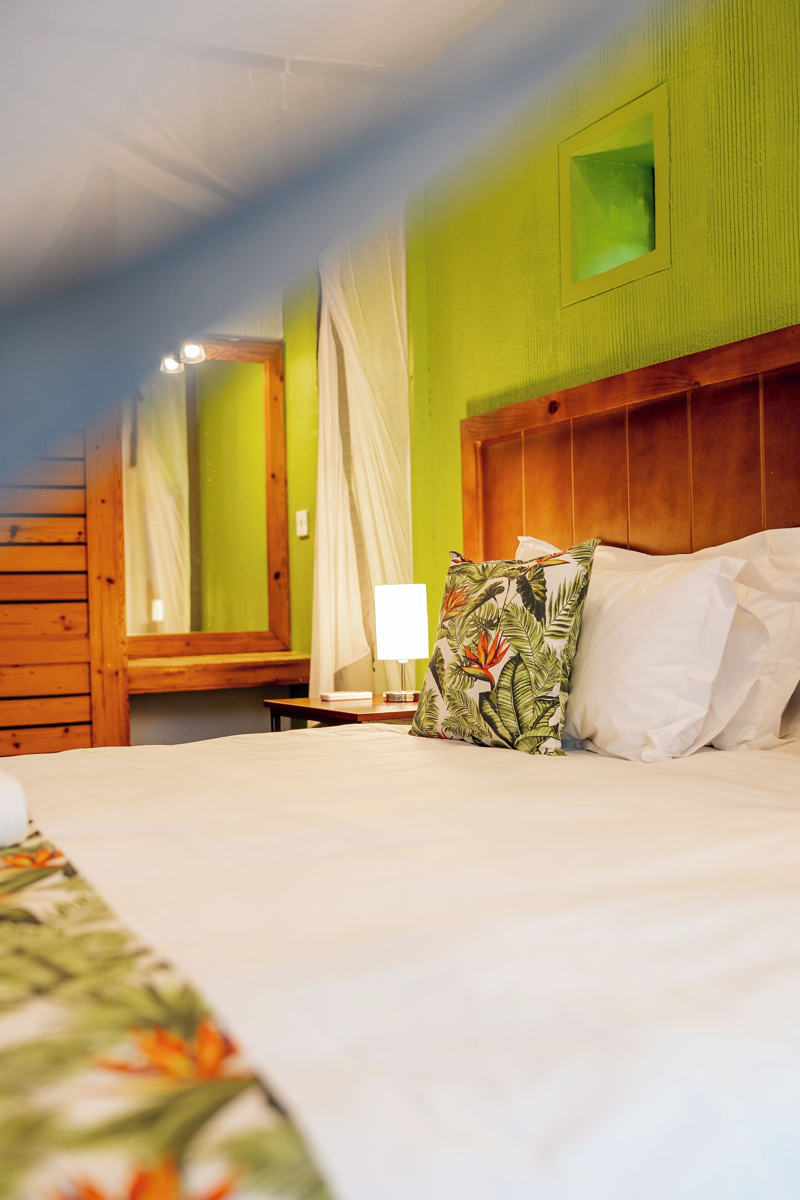 A hotel room with a large bed, a wooden headboard, a nightstand with a lamp, and a window with curtains.