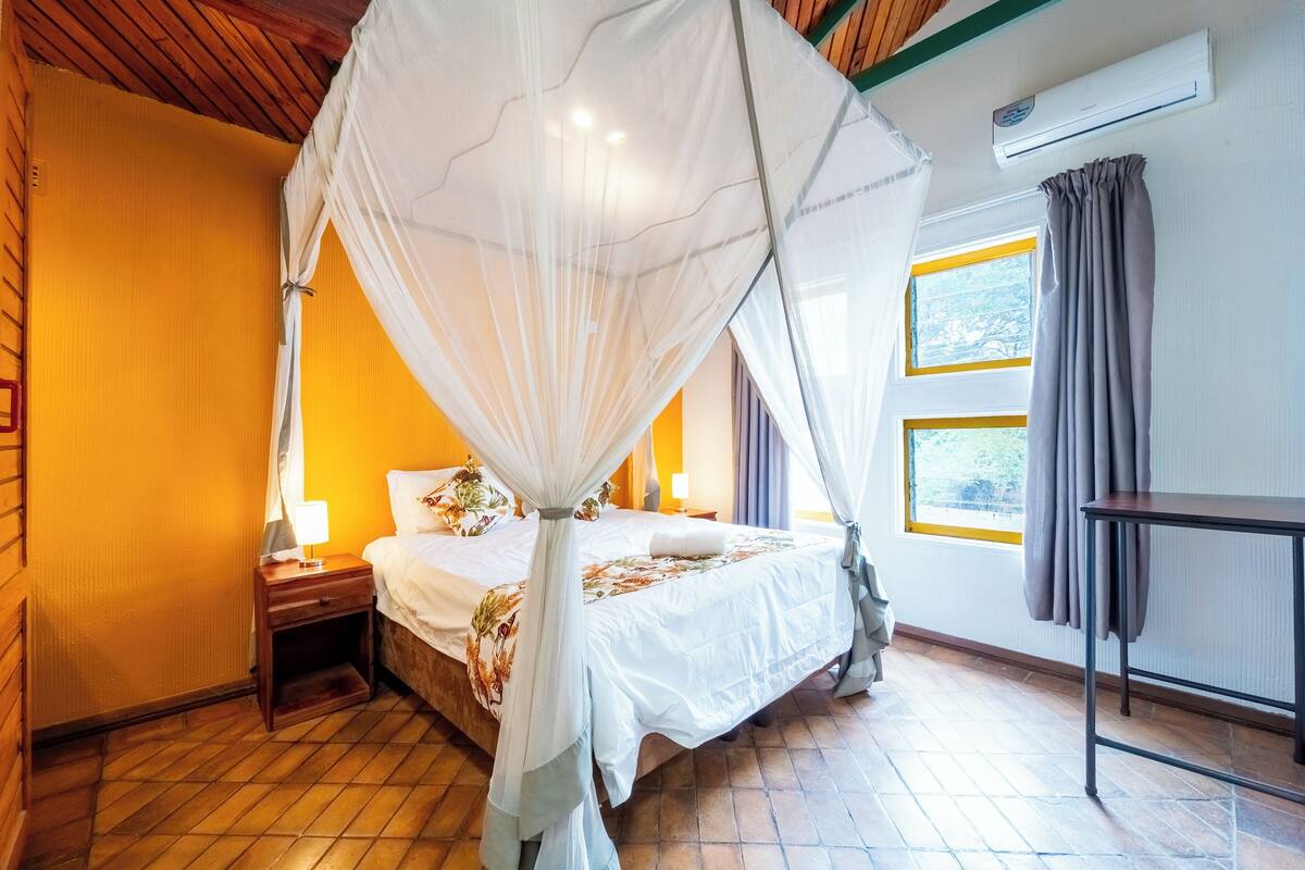 A bedroom with a bed under a canopy, a nightstand, a bedside table, and a window with curtains.