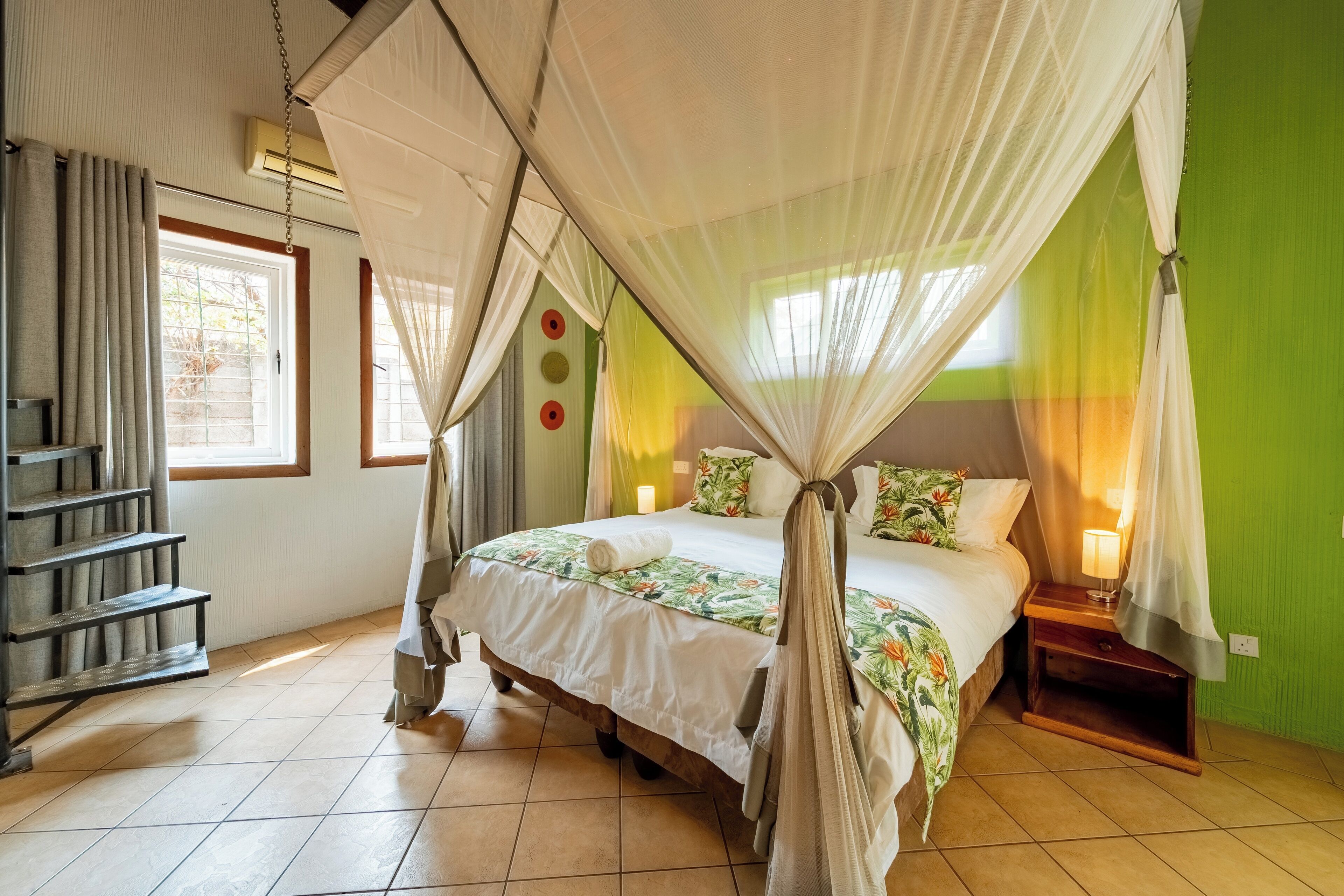 A bedroom with a four-poster bed, a nightstand with a lamp, a wooden staircase, and a green accent wall.