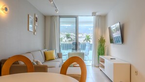 A modern living room with a sofa, dining table, and chairs, a TV mounted on the wall, and a balcony with a view of the city.