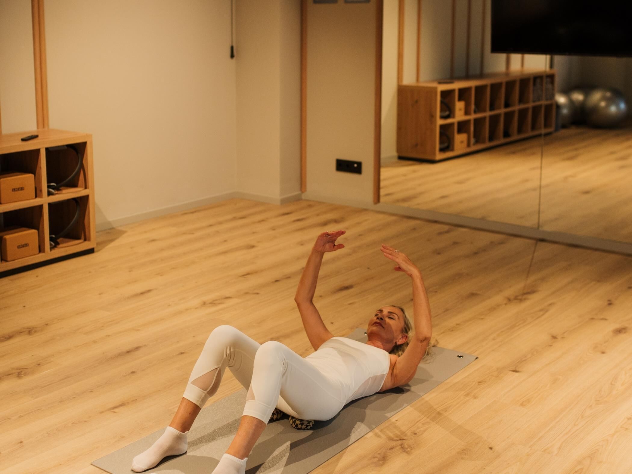 Une personne allongée sur un tapis de yoga dans une pièce dont le sol est en bois, qui contient un miroir et un téléviseur fixé au mur.