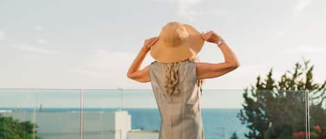 A person standing on a balcony overlooking a body of water, wearing a hat and a sleeveless dress.