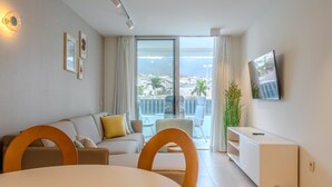 Una sala de estar moderna con sofá, mesa de comedor y sillas, televisor montado en la pared y balcón con vista a la ciudad.