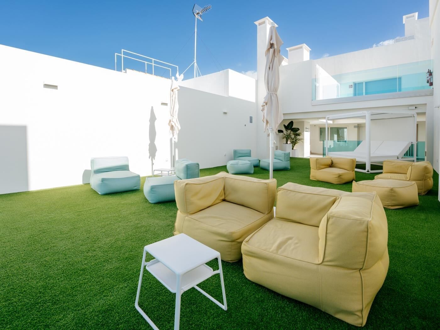 Un espacio lounge exterior moderno con césped artificial verde, asientos color beige y azul claro, y una mesa blanca.